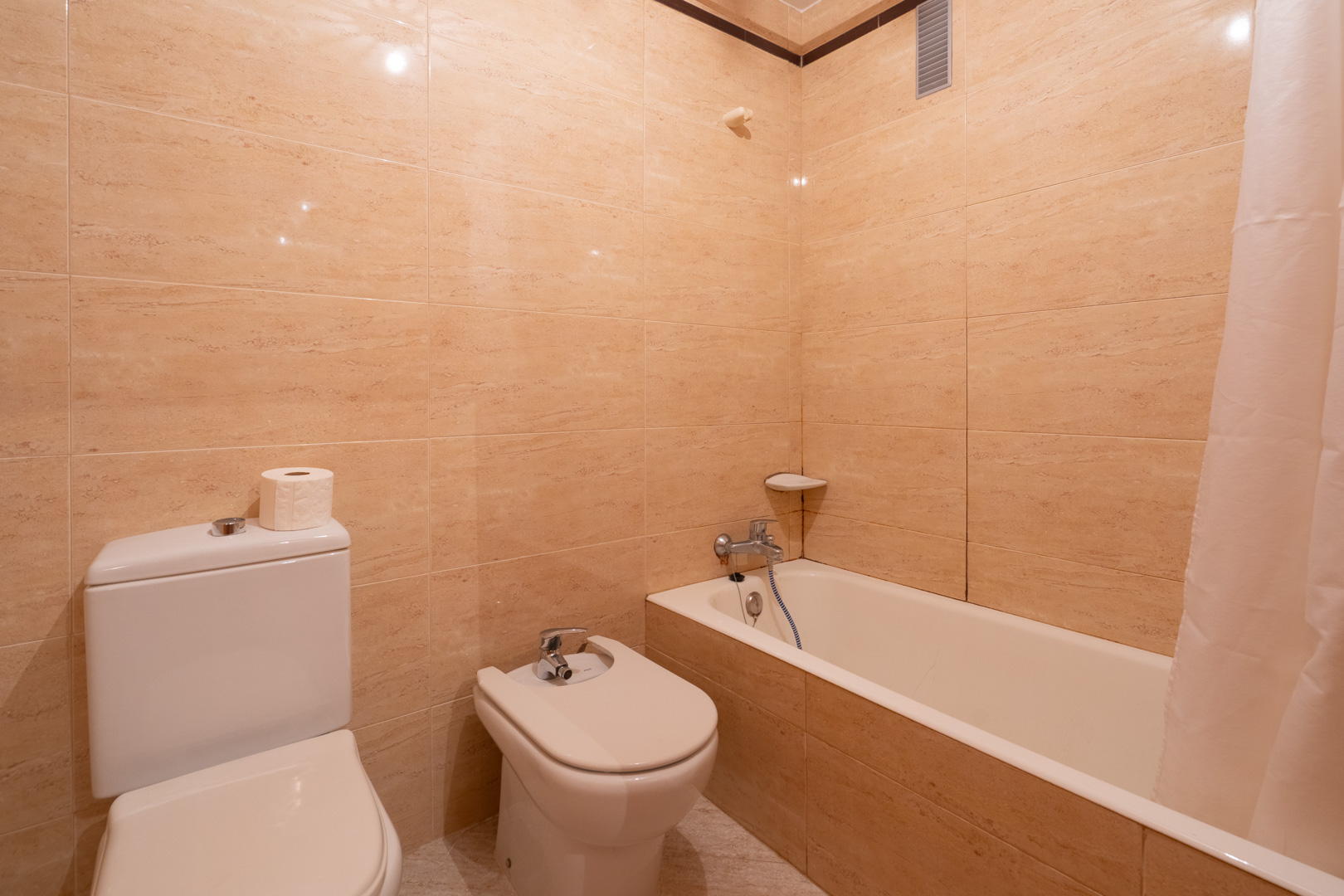 Badezimmer mit beigefliesten Wänden, ausgestattet mit WC, Bidet und Badewanne mit Duschvorhang. Chromarmaturen und eine Eckablage sind vorhanden.