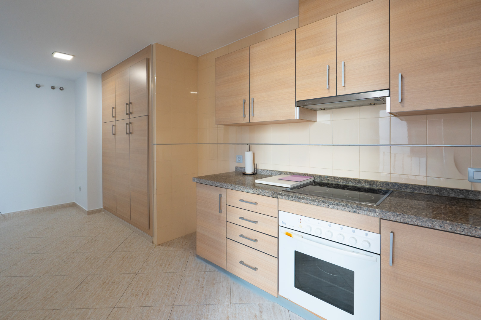 Funktionale Küche mit hellen Holzschränken, Granit-Arbeitsplatte, integriertem Cerankochfeld und Backofen. Hoher Vorratsschrank und beige Bodenfliesen.