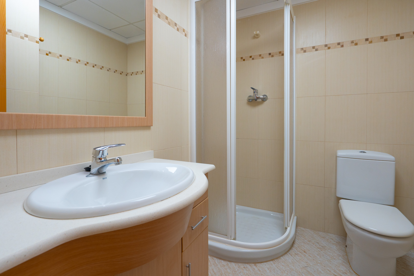 Funktionales Badezimmer mit ovalem Waschbecken auf hellem Holzunterschrank, gerahmtem Spiegel, Eckdusche und beigen Fliesen mit Zierbordüre.