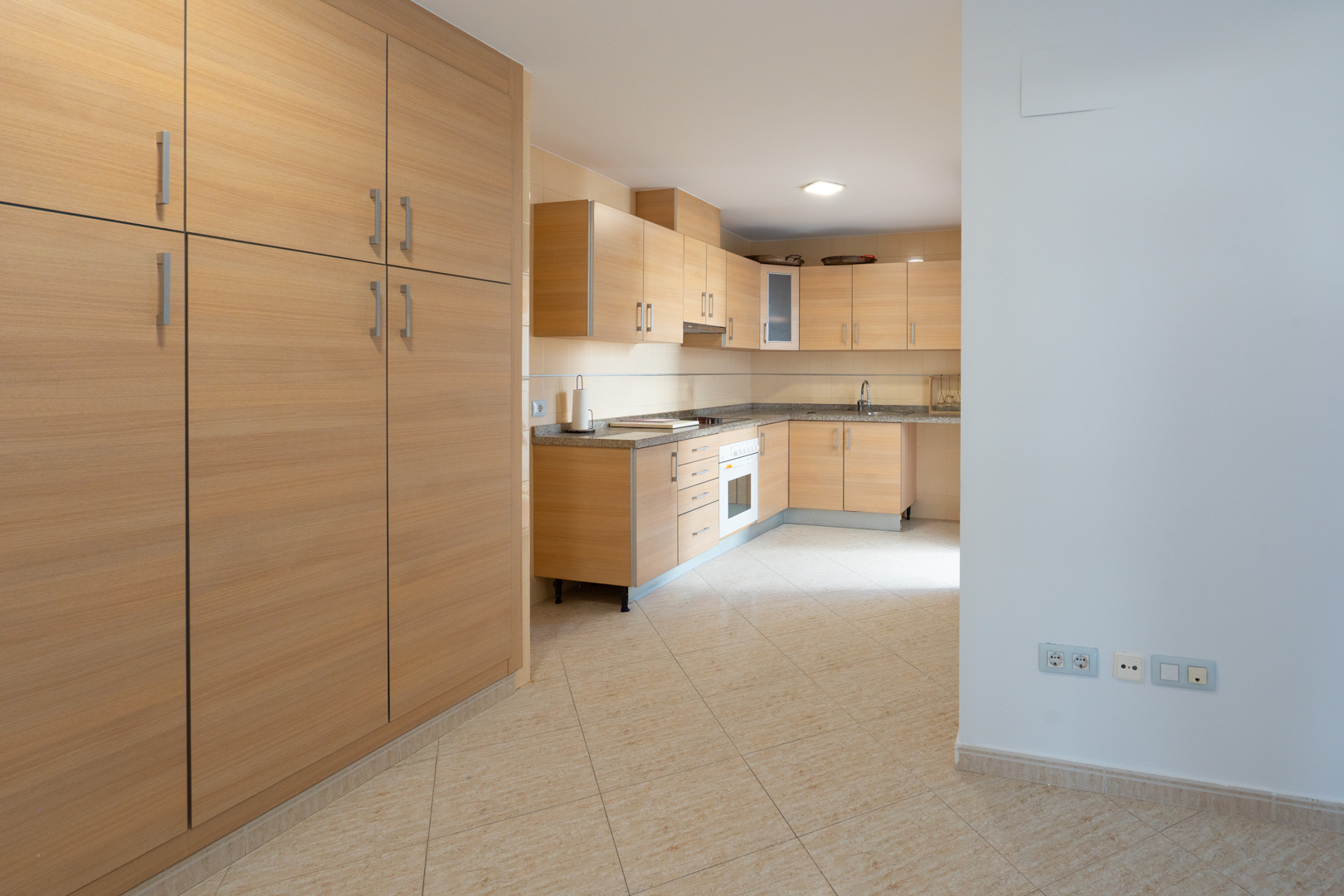 Geräumige Küche mit hellen Holzschränken, gesprenkelten Granitarbeitsplatten und integriertem Backofen. Ein großer passender Schrank steht links auf dem beigefarbenen Fliesenboden.