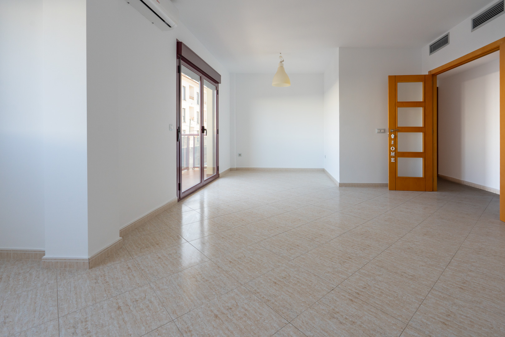 Geräumiger Raum mit beigem Keramikfliesenboden. Große Glasschiebetür (dunkelroter Rahmen) zur Terrasse, Split-Klimaanlage und helle Holztür mit satinierten Glasscheiben sind sichtbar.