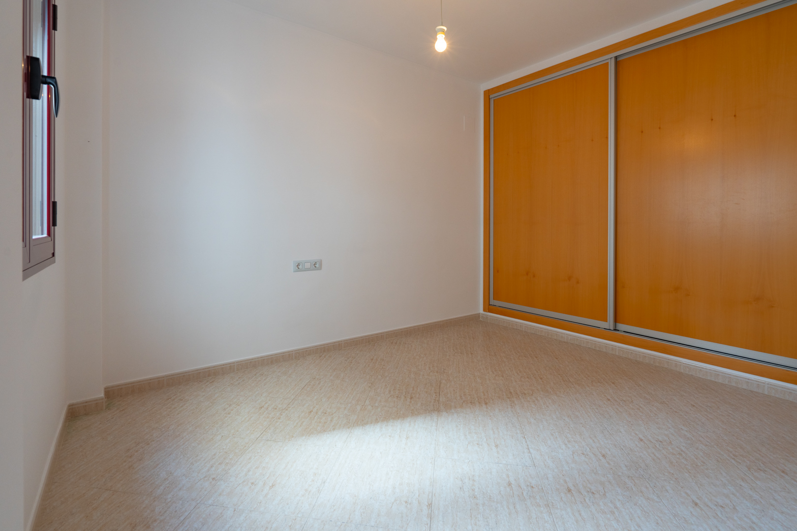 Helles Zimmer mit Einbauschrank, dessen Schiebetüren aus hellem Holz gefertigt sind, und beigefarbenem Fliesenboden. Ein Fenster mit dunklem Rahmen sorgt für Tageslicht.