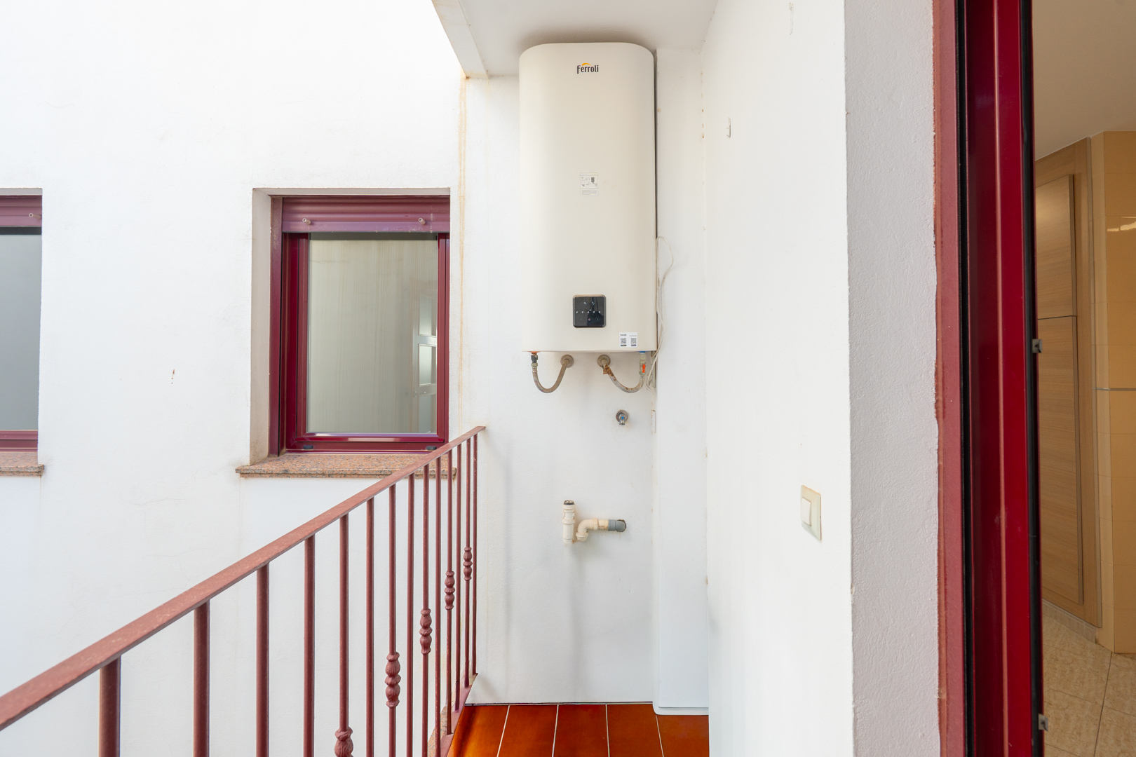 Innenhof mit einem wandmontierten Ferroli-Warmwasserbereiter, Terrakotta-Bodenfliesen und einem roten Metallgeländer. Ein bordeauxrotes Fenster mit Granitfensterbank und passender Türzarge sind sichtbar.