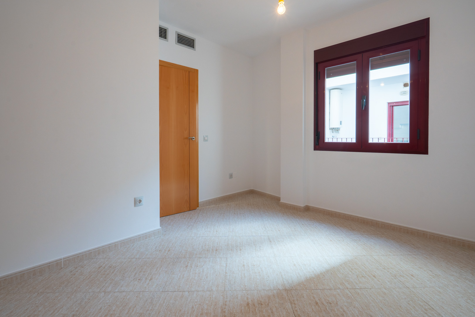 Leerer Raum mit hellen Fliesen und weißen Wänden. Ausgestattet mit einer Holztür und einem burgunderroten Fensterrahmen, blickt es auf einen Innenhof mit Warmwasserbereiter. Lüftungsgitter sind sichtbar.