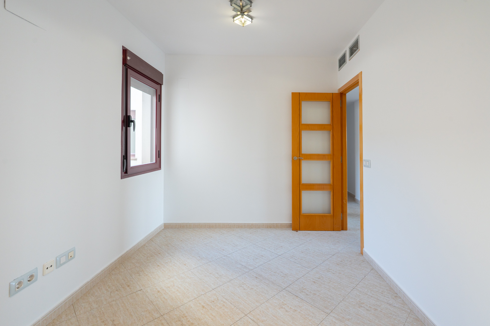 Leerer Raum mit weißen Wänden, bordeauxrotem Fensterrahmen und hellbeigen Diagonalfliesen. Eine Holztür mit Glaseinsätzen und Klimaanlagenlüftung sind sichtbar.