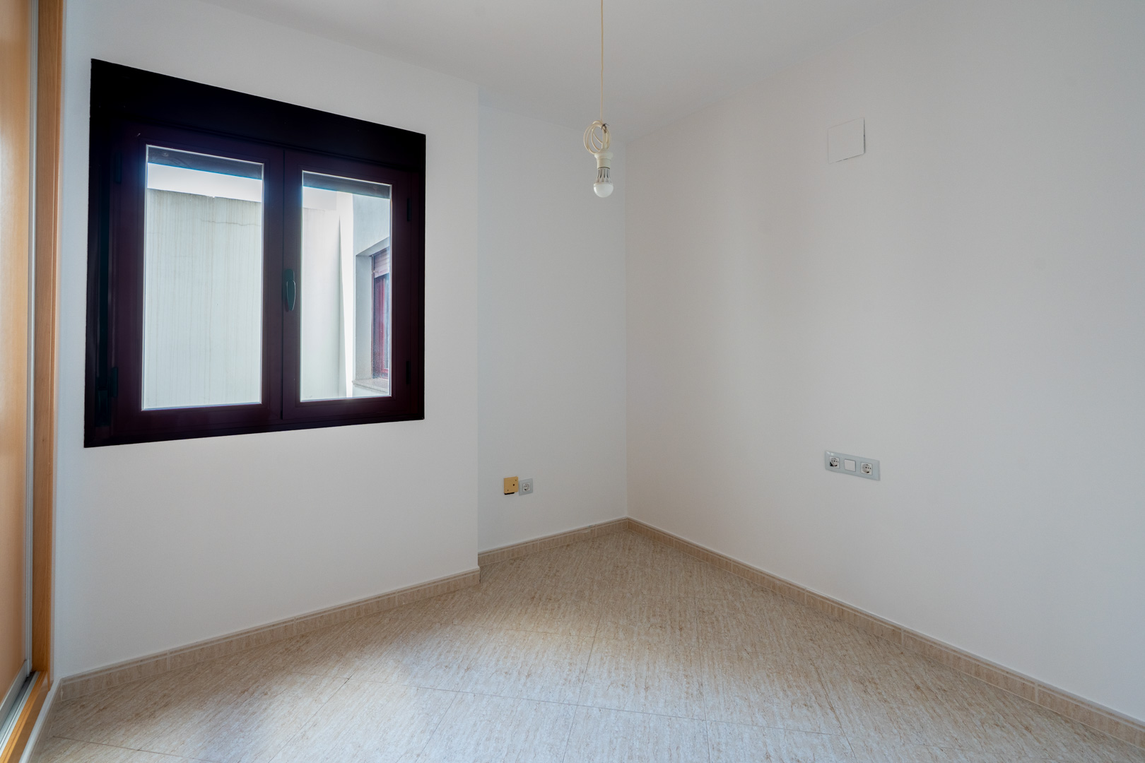 Leerer Raum mit weißen Wänden und hellen Fliesenböden. Das Zimmer verfügt über ein dunkel gerahmtes Fenster zum Innenhof und eine Deckenlampe mit Glühbirne.