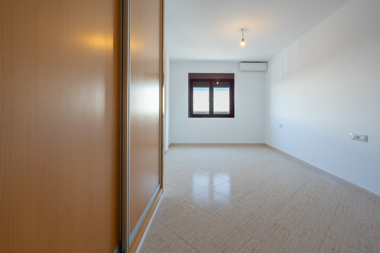 Leeres Zimmer mit hellem Einbauschrank aus Holz, diagonal verlegten beigen Fliesen und Fenster mit Blick auf Dächer. Eine Klimaanlage ist vorhanden.