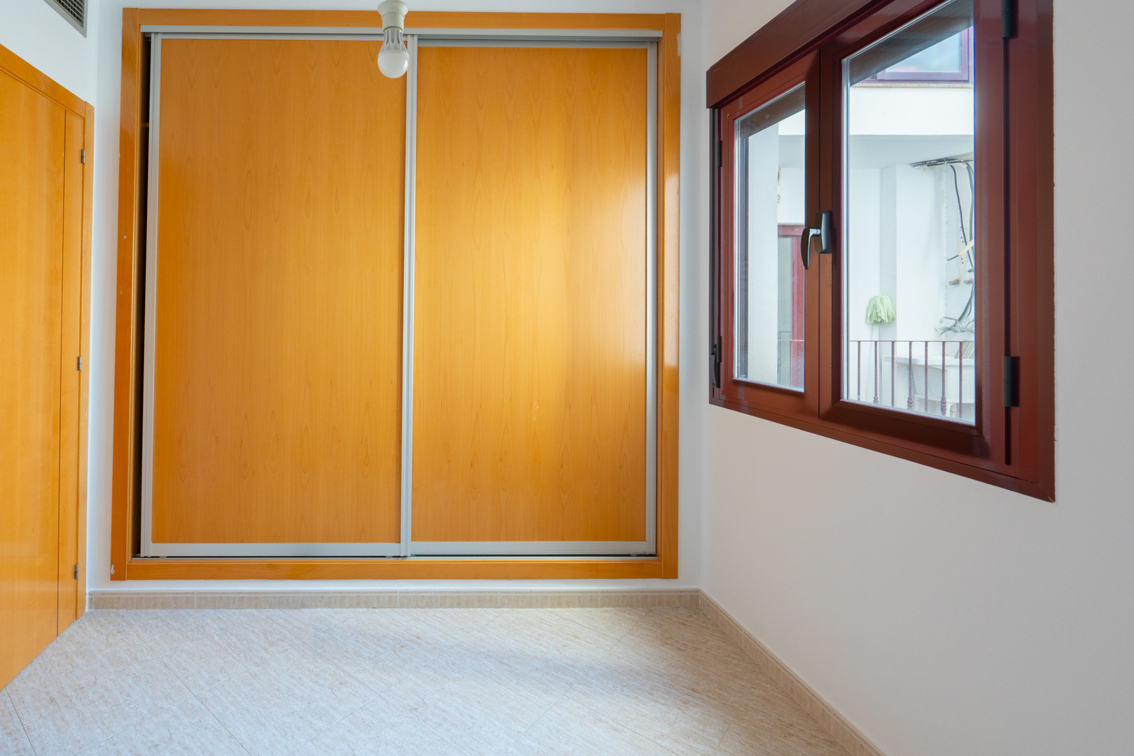 Nebenschlafzimmer mit geräumigem Einbauschrank mit hellen Holzschiebetüren und dunkelbraunem Holzfenster mit Blick auf einen Innenhof.