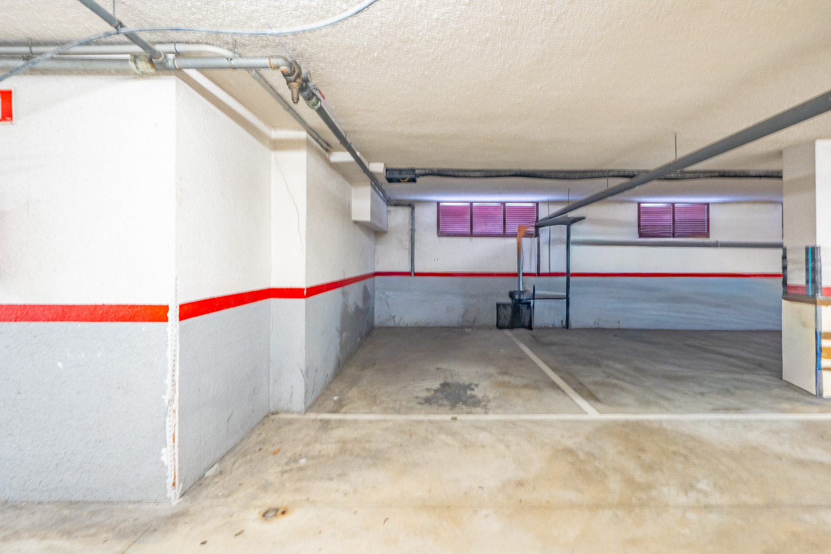 Privater Tiefgaragenstellplatz mit Betonboden, weiß-grauen Wänden mit rotem Streifen und sichtbaren Deckenrohren. Zwei kleine Fenster mit roten Jalousien sind erkennbar.