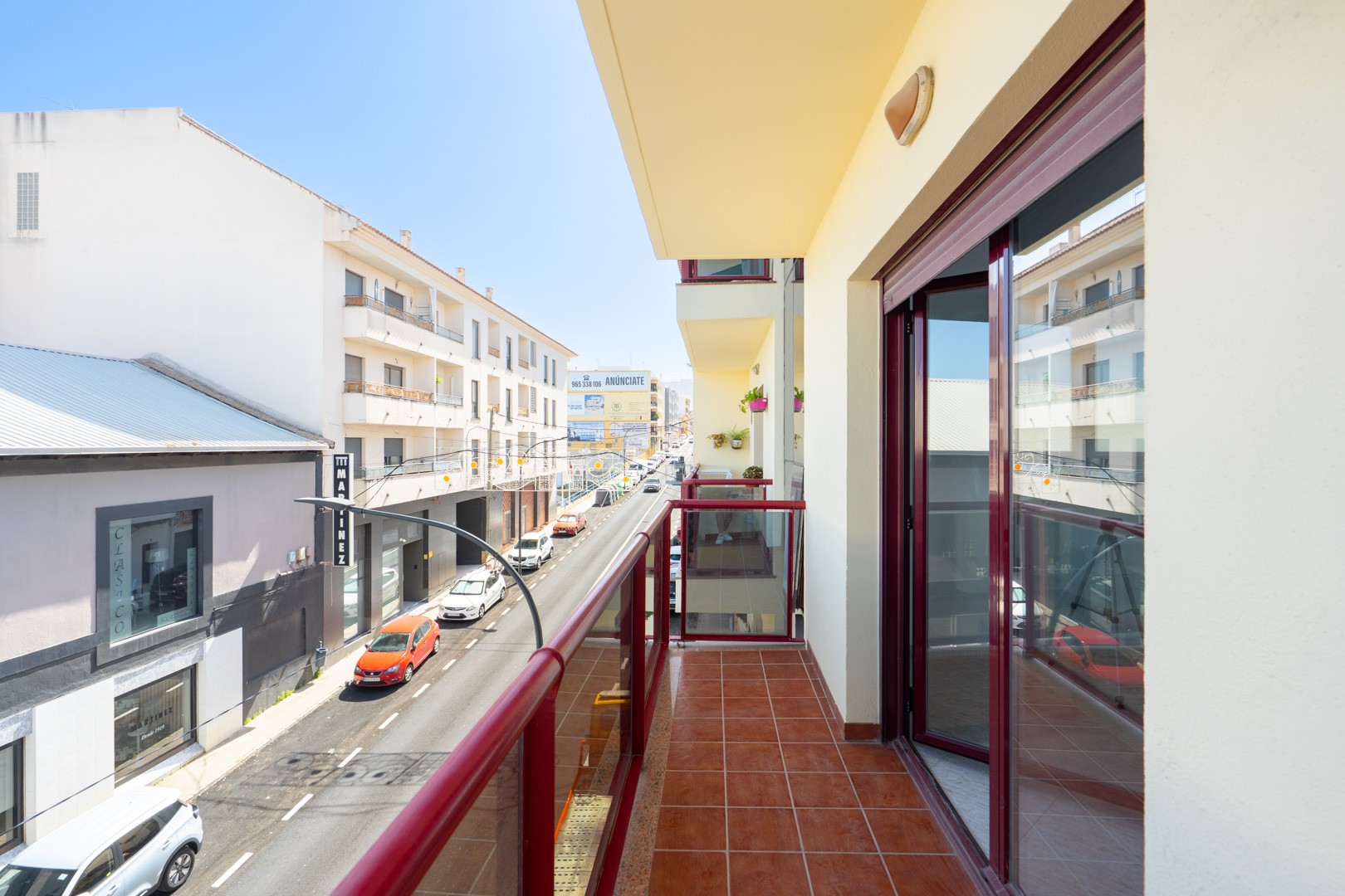 Sonniger Balkon mit Terrakottafliesen und rot gerahmtem Glasgeländer, mit Blick auf eine belebte Stadtstraße und die umliegenden Gebäude.