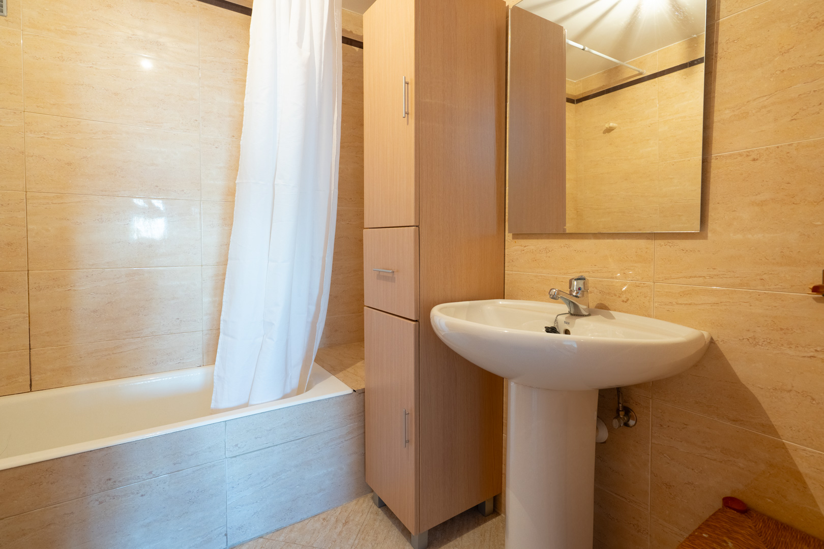Zweites Badezimmer mit weißem Standwaschbecken, hohem Holzschrank und Badewanne mit Duschvorhang. Die Wände sind mit hellbeigen Fliesen verkleidet.