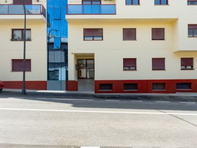 Außenansicht des Wohngebäudes mit cremefarbener Fassade, blauen Glasbalkonen und roten Rollläden. Ein zentraler blauer Glasabschnitt und ein ebenerdiger Eingang mit Rampe prägen das Erscheinungsbild.