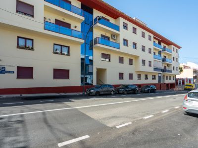 Außenansicht des Wohngebäudes mit heller Fassade, blauen Glasbalkonen, Fenstern mit weinroten Rollläden und einer vertikalen Glasfront.