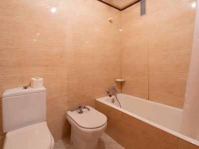 Badezimmer mit beigefliesten Wänden, ausgestattet mit WC, Bidet und Badewanne mit Duschvorhang. Chromarmaturen und eine Eckablage sind vorhanden.