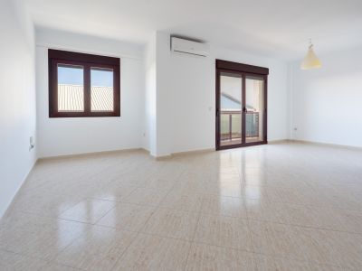 Geräumiger Raum mit hellbeigefliestem Boden, weißen Wänden, einem Fenster und einer Schiebetür mit braunen Rahmen zur Terrasse. Eine Klimaanlage ist installiert.