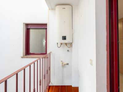 Innenhof mit einem wandmontierten Ferroli-Warmwasserbereiter, Terrakotta-Bodenfliesen und einem roten Metallgeländer. Ein bordeauxrotes Fenster mit Granitfensterbank und passender Türzarge sind sichtbar.