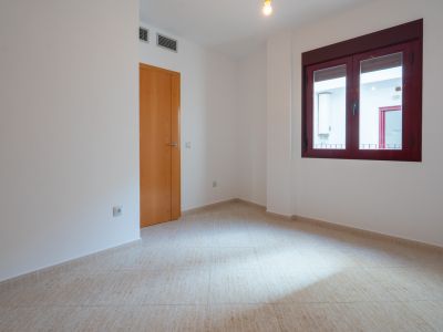 Leerer Raum mit hellen Fliesen und weißen Wänden. Ausgestattet mit einer Holztür und einem burgunderroten Fensterrahmen, blickt es auf einen Innenhof mit Warmwasserbereiter. Lüftungsgitter sind sichtbar.