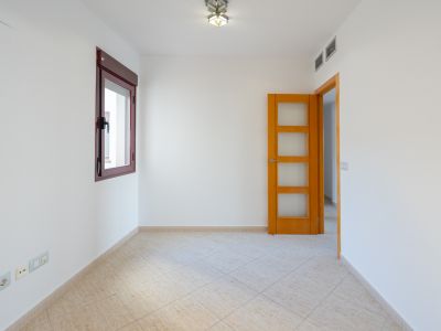 Leerer Raum mit weißen Wänden, bordeauxrotem Fensterrahmen und hellbeigen Diagonalfliesen. Eine Holztür mit Glaseinsätzen und Klimaanlagenlüftung sind sichtbar.