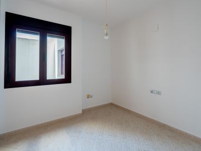 Leerer Raum mit weißen Wänden und hellen Fliesenböden. Das Zimmer verfügt über ein dunkel gerahmtes Fenster zum Innenhof und eine Deckenlampe mit Glühbirne.