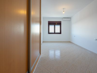 Leeres Zimmer mit hellem Einbauschrank aus Holz, diagonal verlegten beigen Fliesen und Fenster mit Blick auf Dächer. Eine Klimaanlage ist vorhanden.