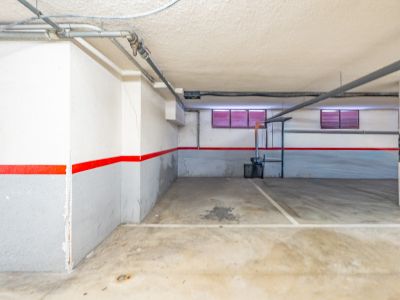 Privater Tiefgaragenstellplatz mit Betonboden, weiß-grauen Wänden mit rotem Streifen und sichtbaren Deckenrohren. Zwei kleine Fenster mit roten Jalousien sind erkennbar.