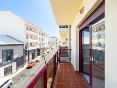 Sonniger Balkon mit Terrakottafliesen und rot gerahmtem Glasgeländer, mit Blick auf eine belebte Stadtstraße und die umliegenden Gebäude.