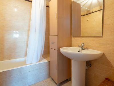 Zweites Badezimmer mit weißem Standwaschbecken, hohem Holzschrank und Badewanne mit Duschvorhang. Die Wände sind mit hellbeigen Fliesen verkleidet.