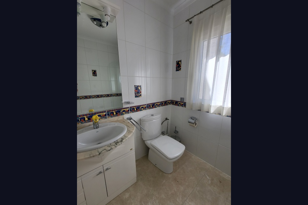 Badezimmer mit weißen Fliesenwänden und dekorativer Bordüre. Ausgestattet mit einem Waschtisch mit heller Marmorplatte, einer Toilette und einem Fenster mit transparenten Vorhängen.