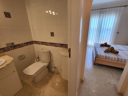 Blick aus einem gefliesten Badezimmer mit Toilette und Waschbecken in ein helles Schlafzimmer mit Einzelbetten, gestreifter Bettwäsche und großem Fenster.