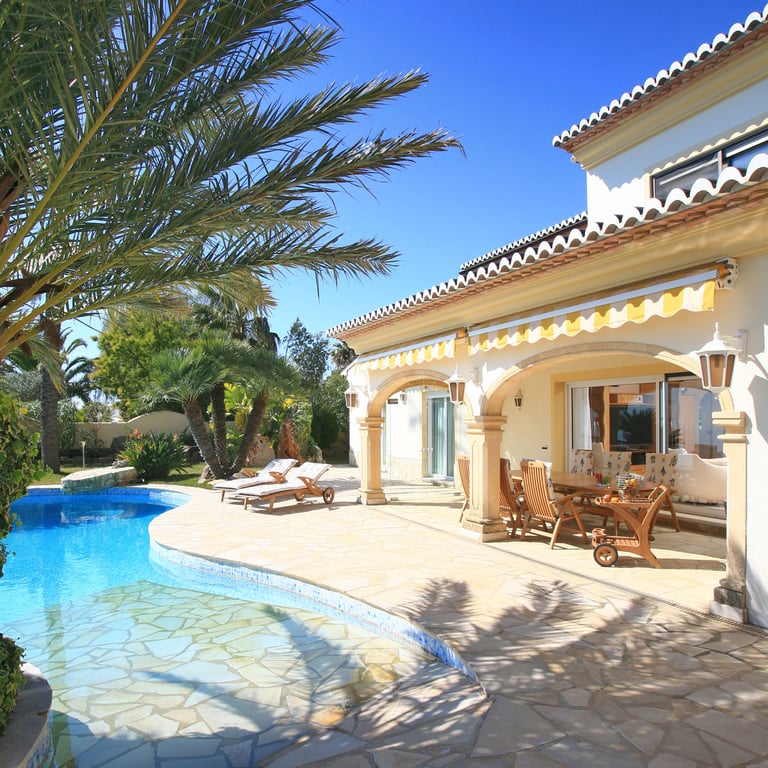 Der Außenbereich der Villa zeigt einen Freiform-Swimmingpool und eine weitläufige Steinterrasse, die zu einer überdachten Naya mit Holz-Essgruppe und gestreiften Markisen führt.