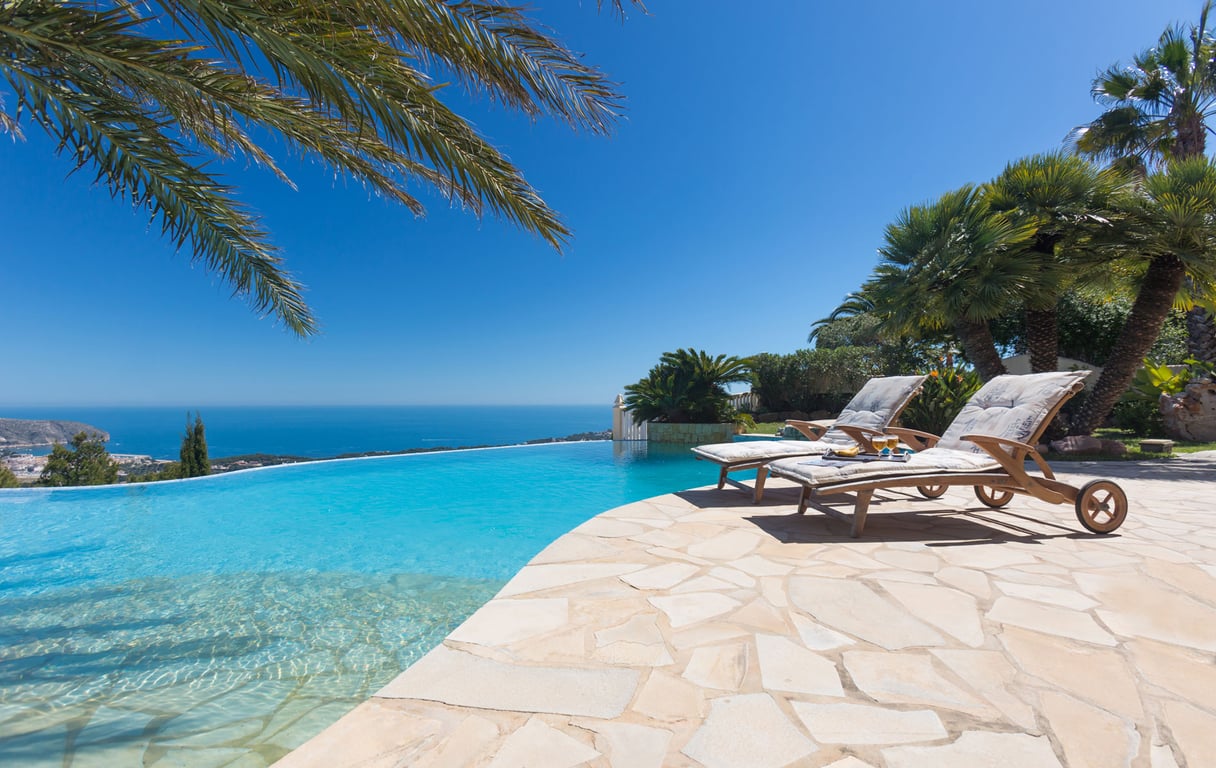 Die Infinity-Poolterrasse mit zwei Holzliegen auf einem Steinplatten-Patio bietet weite Ausblicke auf das azurblaue Mittelmeer und die ferne Küste.