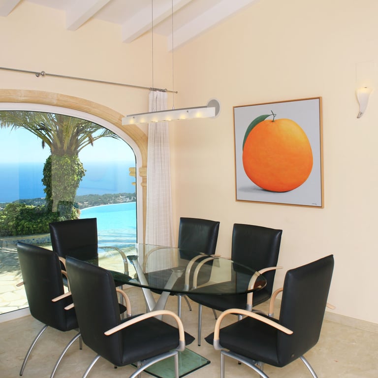 Essbereich mit Glastisch und modernen Stühlen, akzentuiert durch ein großes Rundbogenfenster mit Panoramablick auf Meer und Küste. Ein Kunstwerk mit einer Orange ziert die Wand.