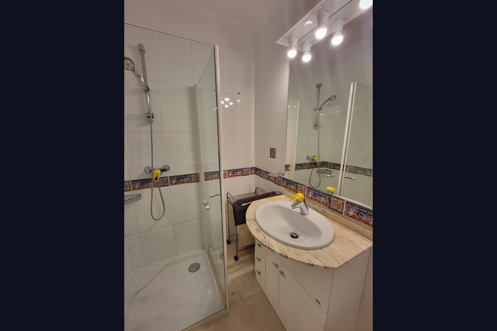 Funktionales Badezimmer mit begehbarer Glasdusche, Waschtisch mit hellem Marmor-Arbeitsplatte und weißem Unterschrank. Weiße Wandfliesen sind mit einem dekorativen mediterranen Bordürenband versehen.