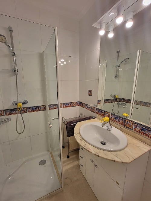 Funktionales Badezimmer mit Glasduschkabine, Waschtisch mit heller Marmorplatte und beleuchtetem Spiegel. Weiße Fliesen mit dekorativer mediterraner Bordüre.