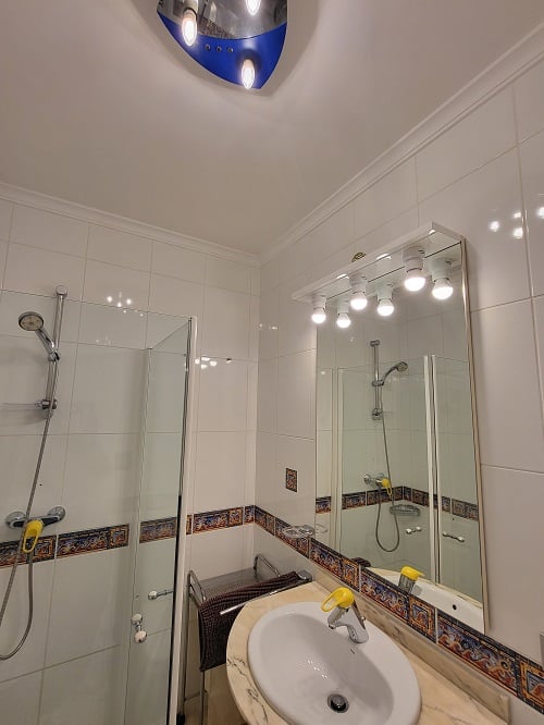 Funktionales Badezimmer mit weißen Fliesen und dekorativer Bordüre, Glasduschkabine und Waschtisch mit Marmor-Effekt-Arbeitsplatte. Beleuchteter Spiegel und blaue Deckenleuchte.