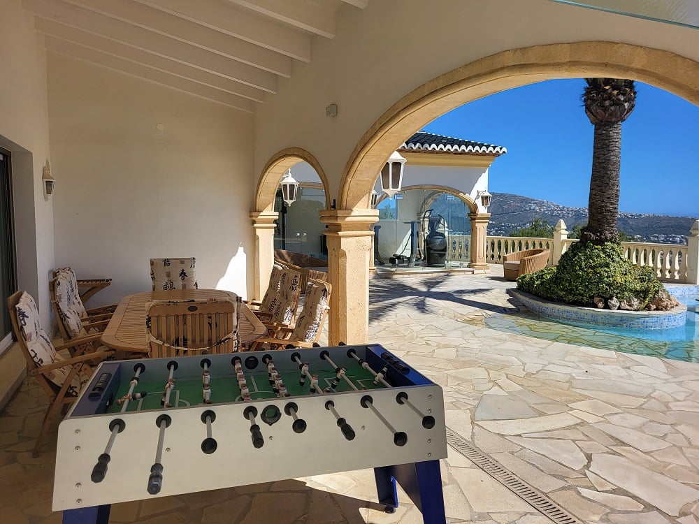 Geräumige überdachte Naya mit Esstisch aus Holz und Kicker, eingerahmt von Steinbögen und unregelmäßigem Steinboden. Bietet Blick auf den Pool mit Palmeninsel und ferne Berge.