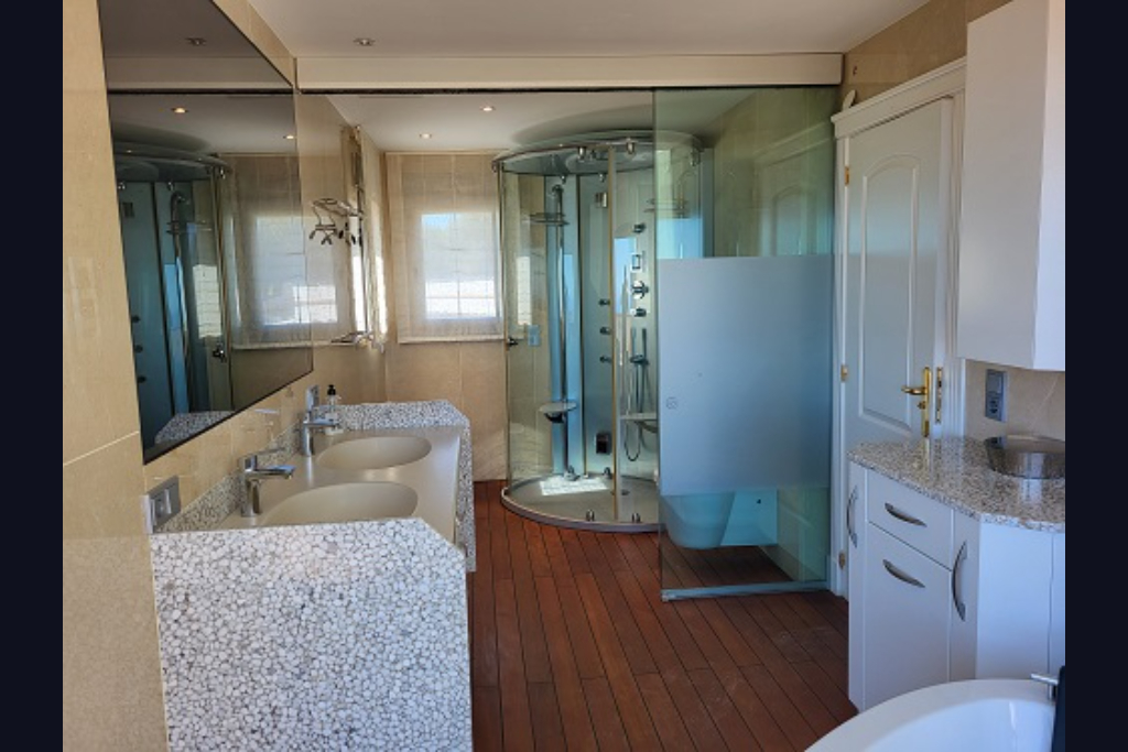 Geräumiges Badezimmer mit Doppelwaschtisch im Kieselstein-Design, runder Hydromassage-Dusche und warmem Holzboden. Großer Spiegel und Fenster mit Jalousien.