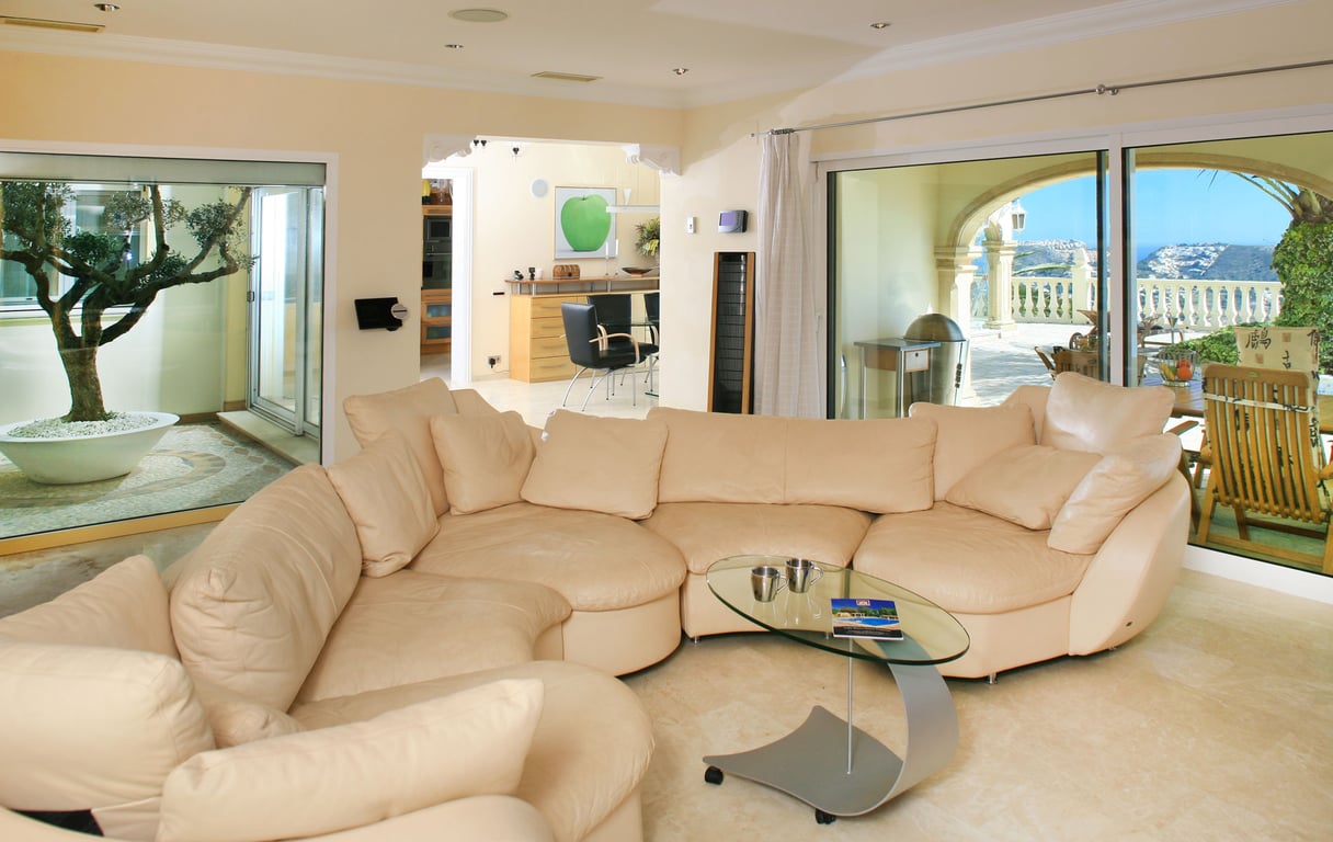 Geräumiges Wohnzimmer mit hellem Ecksofa, Glastisch und Marmorboden. Große Fensterfronten öffnen sich zu einer Meerblickterrasse und einem Innenhof mit Olivenbaum.
