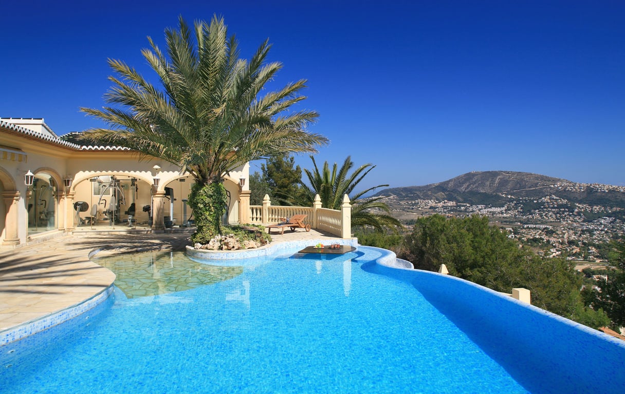 Geschwungener Mosaikpool mit Palmeninsel und Unterwassertisch. Verglaster Fitnessbereich und Terrasse mit Panoramablick auf die Berge.