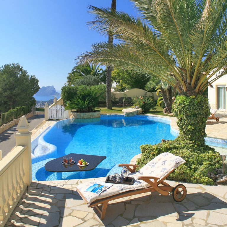 Geschwungener Pool mit einem versenkten schwarzen Tisch und Erfrischungen, angrenzend an eine steingepflasterte Terrasse mit Sonnenliege. Ferne Meerblicke und eine markante Felsformation sind sichtbar.