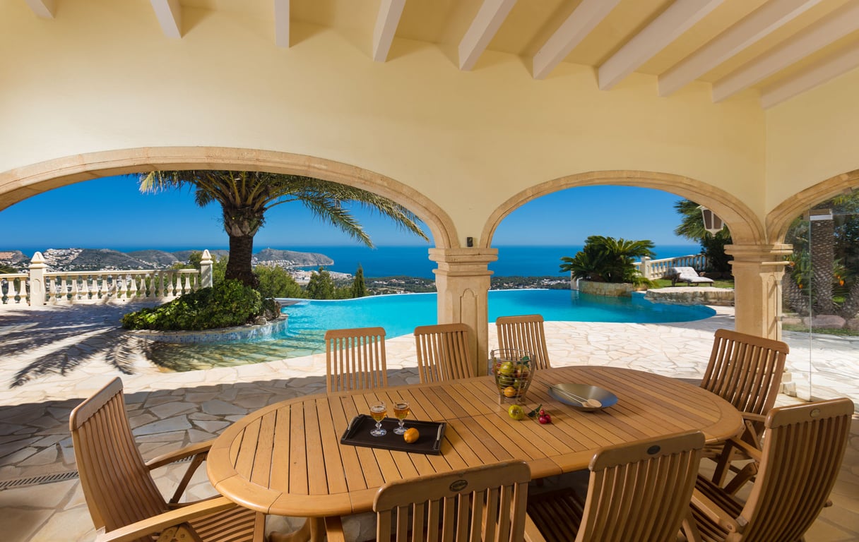 Großzügige überdachte Naya mit Esstisch aus Holz und Stühlen, die durch Steinbögen einen Panoramablick auf das Mittelmeer, die Küste und einen Infinity-Pool freigibt.