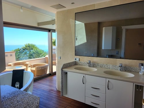 Hauptbadezimmer mit Doppelwaschtisch, Whirlpool-Badewanne und direktem Zugang zu einer Holzterrasse mit Korbmöbeln und Meerblick.