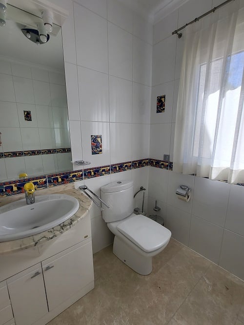 Helles Badezimmer mit weißen Wandfliesen, farbiger Dekorleiste und Akzentstücken. Ausgestattet mit Marmorwaschtisch, integriertem Waschbecken, moderner Toilette und Fenster mit Gardine.