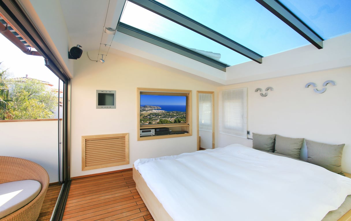 Helles Schlafzimmer mit großem Glasdachfenster und warmem Holzboden. Eine Schiebetür führt zur privaten Terrasse mit Korbsessel; ein eingebauter TV zeigt Küstenblicke.