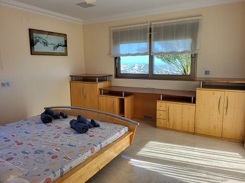Helles Schlafzimmer mit hellem Holzdoppelbett, integriertem Schreibtisch und Schränken. Die Fenster bieten Panoramablicke auf die Küste und füllen den Raum mit Tageslicht.