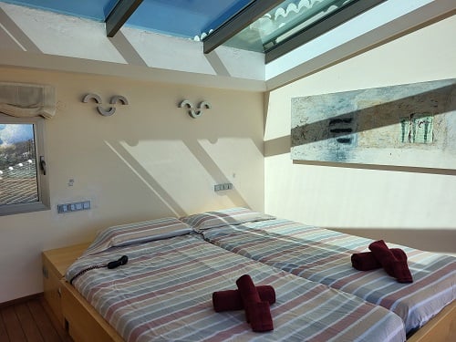 Helles Schlafzimmer mit zwei Einzelbetten, gestreifter Bettwäsche und roten Handtüchern. Ein großes Dachfenster sorgt für natürliches Licht, ergänzt durch ein abstraktes Gemälde an der Wand.