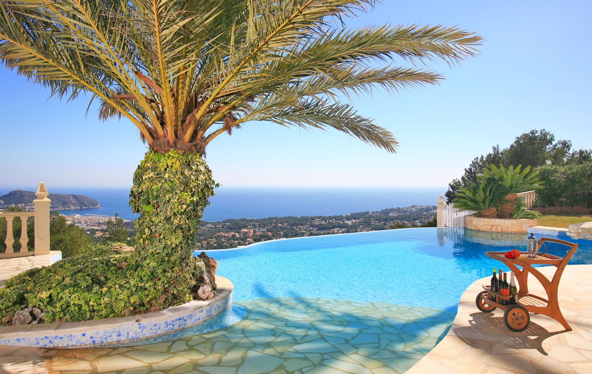 Infinity-Pool mit Panoramablick auf Meer und Küste, inklusive einer von Efeu bewachsenen Palme und einem Holz-Servierwagen auf der Steinterrasse.