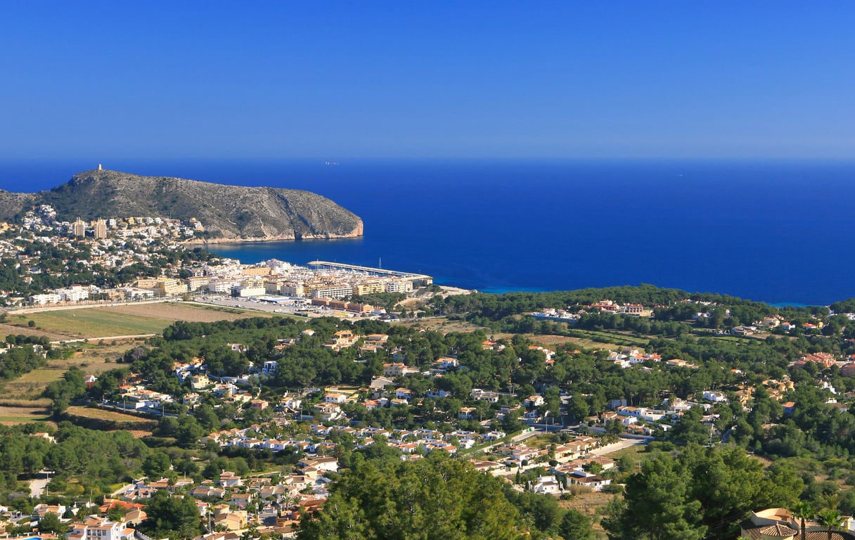 Panoramablick von einem Hügel, mit einer Küstenstadt, einem Yachthafen, einem markanten Berg und dem tiefblauen Mittelmeer unter klarem Himmel.