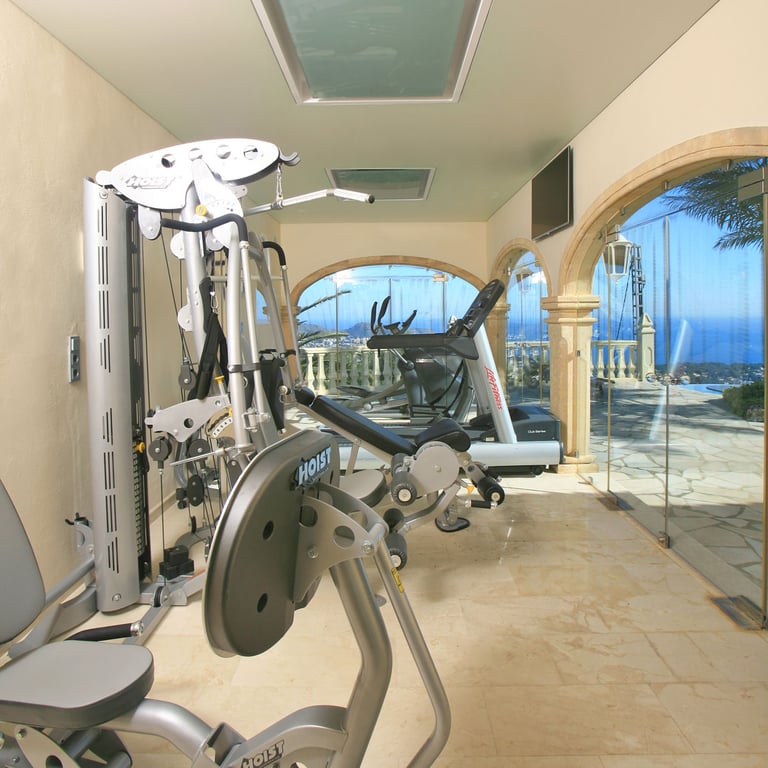 Privater Fitnessraum mit Hoist- und Life Fitness-Geräten, Marmorboden und gewölbten Glaswänden mit Panoramablick auf Meer und Pool. Dachfenster sorgen für Tageslicht.