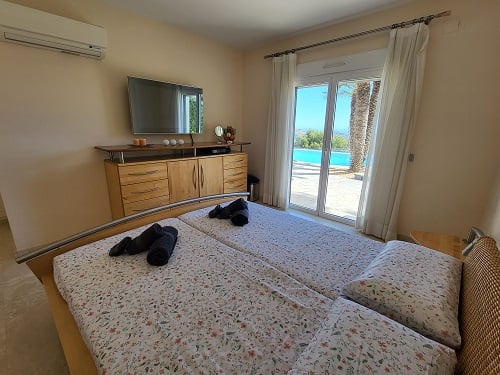 Schlafzimmer mit Einzelbetten, Wandfernseher und direktem Terrassenzugang mit Blick auf Pool und Palmen. Klimaanlage und Holzmöbel.